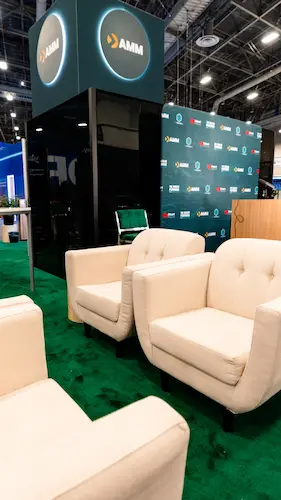 Custom trade show exhibit lounge with plush white armchairs, artificial turf carpeting, and a modern illuminated AMM display tower, built by SoftSpace Exhibits.