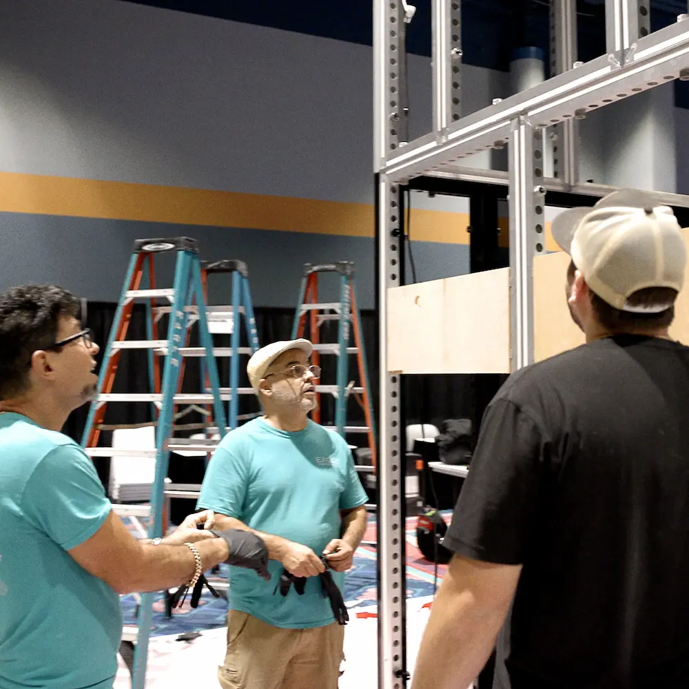 Three SoftSpace team members constructing an aluminum exhibit booth framework on a trade show floor, representing end-to-end sketch, build, and teardown show services.